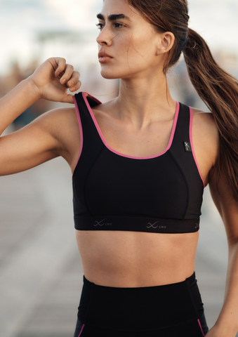 A woman wearing a black sports bra with pink trim stands outdoors with one arm raised, looking to the side. She appears to be in an athletic or fitness setting. The background is blurred.