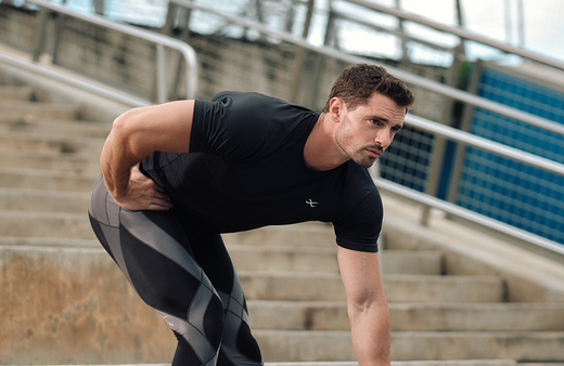 A man wearing a black athletic shirt and patterned leggings stretches his right leg on outdoor concrete steps, reaching down to touch his right foot with his right hand. A metal railing and blue sky are in the background.