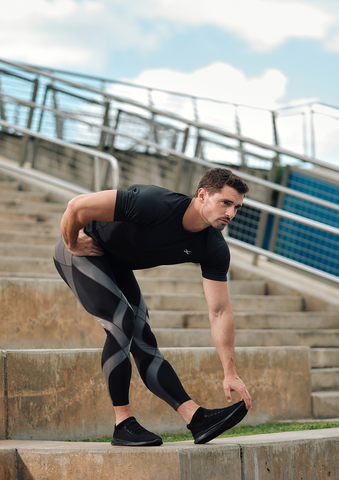 A man wearing a black athletic shirt and patterned leggings stretches his right leg on outdoor concrete steps, reaching down to touch his right foot with his right hand. A metal railing and blue sky are in the background.