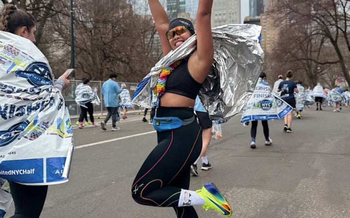 A runner wearing athletic gear, sunglasses, and a finisher's foil blanket jumps with arms raised on a city street after a race. Other participants with foil blankets are visible in the background.