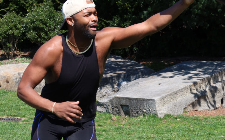 A man wearing a backward cap, sleeveless black shirt, and black shorts stands on grass with one arm extended and the other bent, appearing to throw or catch something outdoors.