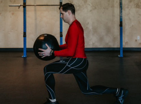 A man in athletic clothing performs a lunge while holding a medicine ball with both hands in a gym with a barbell rack in the background.