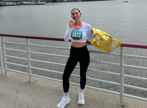 A person wearing a race bib numbered 3072 stands by a waterfront railing, smiling and holding a medal to their mouth with one hand and a yellow blanket with the other. The background shows water and distant buildings.