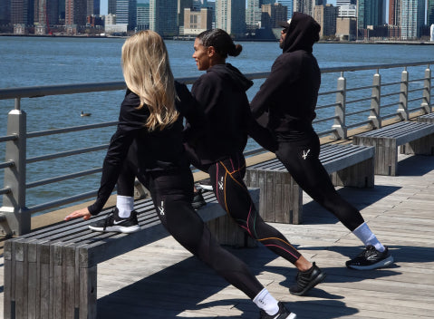 Three people in athletic wear stretch their legs on a wooden pier next to a metal railing by the water, with city buildings visible across the river in the background.